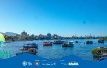 Fotografia horizontal em plano aberto. Uma vista panorâmica do Rio Itiberê em um dia ensolarado. O céu, no terço superior, é de um azul intenso, sem nuvens. A água do rio, em tons de azul e verde escuro, preenche o primeiro plano e o centro. Diversas embarcações de pequeno e médio porte flutuam na água, distribuídas horizontalmente. Em primeiro plano, à direita, está um barco de madeira com o casco verde-escuro parcialmente oculto pela faixa azul da sobreposição. No centro, à esquerda, destaca-se um barco de pesca tradicional com toldo amarelo e casco escuro. Ao fundo, a linha do horizonte exibe a silhueta da cidade de Paranaguá: prédios, guindastes do porto e vegetação rasteira. Um morro com densa mata verde limita a paisagem à direita. A iluminação é forte, criando reflexos brilhantes na superfície da água. Na base, há uma faixa azul ondulada com o logo do projeto e os créditos de patrocínio.