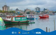 Fotografia em plano aberto, capturando a paisagem urbana portuária de Paranaguá em um dia nublado. Em primeiro plano, a água da baía é calma e cinzenta. Três grandes barcos de passageiros dominam o meio-plano. À esquerda, um barco de madeira com o casco verde e o convés branco e laranja, com o nome "Swell" pintado na lateral, está em destaque. Ao seu lado, um barco azul e branco com toldo azul flutua, e à direita, um barco de casco vermelho vibrante com detalhes brancos ancora. Entre o barco verde e o azul, um homem em uma pequena canoa branca manuseia um remo. O fundo da imagem é preenchido pelo horizonte de Paranaguá: edifícios modernos mais escuros e altos se misturam com casarios históricos coloridos (tons de rosa, azul e amarelo) da orla. Um cais ou estruturas de madeira são visíveis à extrema direita. O céu está coberto por nuvens cinzentas e carregadas, mas a cena é rica em detalhes da vida marítima e urbana da cidade. No rodapé, a faixa azul do projeto "Pá na Água" com logotipos.
