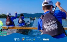 Fotografia horizontal e externa, capturada na altura da água. 3 jovens remam canoas no mar, sob sol forte. O remador em primeiro plano, à direita, está de costas. Ele veste boné branco, camisa azul e colete salva-vidas preto com a estampa "Passaúna Paddle Club". O colete ocupa grande parte do terço direito. O jovem segura o remo com ambas as mãos, preparando a braçada direita. Atrás dele, mais 2 jovens, também de coletes, estão em outra canoa. Eles também remam, impulsionando a canoa escura. A água do mar está calma e reflete o céu azul. À esquerda no primeiro plano, aparece a ponta de um remo verde e amarelo. Em segundo plano, uma canoa de pesca tradicional, de cor amarela com detalhes em madeira, flutua em águas mais distantes. No fundo, a linha do horizonte revela uma área de mata densa e morros. Iluminação natural do dia é intensa, criando uma atmosfera de energia e atividade ao ar livre. Na parte inferior, uma faixa azul com a logo do projeto "Pá na Água" e logos de patrocinadores.