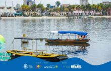 Fotografia horizontal da orla de Paranaguá, sob céu nublado. A água do mar, de tonalidade cinzenta, ocupa a maior parte do primeiro e segundo planos. Duas embarcações de pequeno porte flutuam na água calma. A canoa em primeiro plano, à esquerda, é preta e amarela, com uma estrutura de metal preto coberta por uma lona escura. A outra embarcação, mais ao centro, é colorida — tem casco vermelho e amarelo — e possui um toldo azul claro e escuro, sustentado por estrutura metálica. Um colete salva-vidas laranja pende da lateral desta segunda canoa. [PAUSA BREVE] A linha de costa, em segundo plano, é marcada por uma faixa de areia clara. Acima da areia, a paisagem urbana se estende. Há uma sequência de prédios históricos coloridos e edifícios modernos, com palmeiras altas em destaque. À esquerda, barracas de feira com telhados brancos pontiagudos são visíveis. [PAUSA BREVE] O horizonte apresenta edifícios altos. Na parte inferior, uma faixa azul contém os logos e o título "Pá na Água". A atmosfera é tranquila.