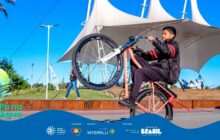 Fotografia horizontal e externa, com foco em um jovem fazendo uma manobra de bicicleta em um espaço público. O jovem, no centro da composição, veste um agasalho preto com detalhes em vermelho e empina uma bicicleta BMX, mantendo a roda dianteira erguida. Ele tem a pele morena e os braços flexionados no guidão. A bicicleta tem a estrutura principal em tom de salmão ou rosa claro. [PAUSA BREVE] Em segundo plano, uma ampla estrutura de lona branca, sustentada por mastros, funciona como pano de fundo. À direita e mais ao fundo, há degraus de madeira onde pessoas estão sentadas ou conversando. [PAUSA BREVE] À esquerda, no meio-plano, uma pessoa caminha na sombra, em frente a coqueiros e vegetação verde. O chão é cinza e plano. O céu é azul e claro. A iluminação é forte e direta, criando sombras nítidas. A cena é dinâmica e transmite a energia do esporte e da cultura urbana. Na parte inferior, uma faixa azul contém os logos e o título "Pá na Água".