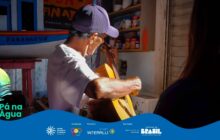 Fotografia horizontal, capturada em ambiente interno com luz quente e contrastada. Um homem de pele bronzeada, no centro, toca um instrumento de cordas de cor amarela, provavelmente um violão. Ele está de perfil, voltado para a esquerda, usando uma camiseta branca de manga curta e um boné roxo escuro. Sua concentração é visível. [PAUSA BREVE] A luz solar intensa ilumina o braço musculoso e o instrumento, criando sombras fortes. À extrema direita, em primeiro plano, uma silhueta de cabelo escuro, provavelmente outra pessoa, corta o enquadramento verticalmente. [PAUSA BREVE] Ao fundo, à esquerda, parte de uma pintura de embarcação branca com a palavra "PARANAGUÁ" em vermelho e azul, decora a parede. Acima da pintura, uma prateleira de madeira simples exibe pequenos objetos coloridos. [PAUSA BREVE] O ambiente é simples, doméstico ou de um pequeno comércio ribeirinho. A cena transmite tranquilidade e a valorização da cultura musical local. Na parte inferior, uma faixa azul contém logos e o título "Pá na Água".