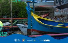 Fotografia horizontal em close-up focado na proa de barcos coloridos atracados na margem. Em primeiro plano, no centro e à direita, destaca-se a proa de uma canoa de madeira pintada em azul vibrante e amarelo forte. Na ponta da proa, uma escultura em madeira azul, que se assemelha a um pássaro ou dragão, funciona como ornamento. A lateral do barco exibe a inscrição "PGUA 421/5543103" em branco. A borda inferior da proa é vermelha. À esquerda, parcialmente visível, está uma segunda embarcação branca chamada "DONA", com detalhes vermelhos. Mais à esquerda, uma terceira embarcação verde e vermelha é parcialmente vista. Ao fundo, um aglomerado de cascos de barcos de cores variadas e uma parede escura de tijolos à direita, todos fora de foco. Folhagens verdes densas ocupam o canto superior esquerdo. A iluminação é natural, destacando as cores e as texturas da madeira. Na parte inferior, uma faixa azul contém os logos do projeto "Pá na Água" e apoiadores.