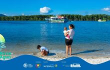 Fotografia horizontal, plano aberto em dia ensolarado. A cena se passa em uma praia de rio ou baía. No primeiro plano, uma mulher jovem, em pé, de costas, segura um bebê no colo. Ela veste camiseta branca e shorts pretos, e está descalça com os pés na areia úmida. O bebê usa roupa branca e está virado de frente para a água. À frente da mulher, uma criança, também de camiseta branca e shorts escuros, está agachada brincando na areia. A areia é clara e forma uma pequena faixa. O segundo plano é dominado pela água azul-escura e vibrante. Ao centro, flutua um grande barco de passageiros, branco com detalhes em vermelho e verde, sob uma nuvem branca solitária no céu azul. No fundo, uma densa faixa de mata verde escura, típica da mata atlântica, preenche o horizonte. À direita, um pequeno barco de pesca amarelo e verde está atracado. Na base, há uma faixa azul com os créditos do projeto.