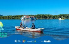 Fotografia horizontal em plano geral, capturando o Rio Itiberê sob sol forte. Em primeiro plano, uma pequena canoa de pesca de madeira, com casco vermelho e interior branco, é remada por duas pessoas. O homem, à esquerda, usa um boné vermelho e uma camiseta escura, remando com vigor. A mulher, sentada na popa à direita, veste roupas escuras e observa o horizonte. A canoa está no terço inferior, sobre a água azul escura e ondulada. Ao fundo, no centro, duas embarcações maiores de pesca estão atracadas: uma branca, vermelha e azul de dois andares, e uma menor, branca e verde, mais à direita. Atrás das embarcações, uma longa faixa de vegetação verde escura e densa, característica de manguezal, ocupa o horizonte. O céu é azul claro e sem nuvens. A luz é intensa, criando um forte contraste. Na parte inferior, uma faixa azul contém os logos do projeto "Pá na Água" e seus apoiadores.