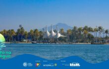 Fotografia horizontal em plano geral, capturando uma orla sob céu claro. Em primeiro plano, a água do mar ou rio é azul escura e levemente ondulada. Acima da água, em segundo plano, domina uma linha horizontal de palmeiras, com folhas verdes e amarelas. No centro, por trás das palmeiras, ergue-se uma estrutura arquitetônica moderna com três coberturas brancas, altas e pontiagudas, que se assemelham a velas de barco infladas. Embaixo das palmeiras e da estrutura, vê-se uma área de calçada e pequenos barcos atracados, como um pequeno cais ou porto. Em terceiro plano, sob o céu azul intenso, uma montanha com pico suave se eleva, dominando o centro superior. A iluminação é forte, de dia ensolarado, destacando as cores vibrantes. À esquerda, parte do logo do projeto "Pá na Água" é visível. A tarja azul com logotipos dos apoiadores está na base.