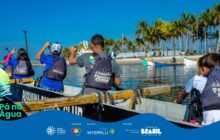 Fotografia horizontal em plano médio-geral, capturada de dentro de uma canoa grande de cor cinza, mostrando 4 jovens remando. Os jovens estão de costas, usando coletes salva-vidas e mochilas escuras com o logo "Passando Paddle Club". Da esquerda para a direita: uma jovem de cabelo preto e dois jovens de boné, sendo o central de camiseta cinza e o da esquerda de azul. O jovem mais à direita é menor e está curvado sobre o remo. Eles seguram os remos e estão na atividade de canoagem. O sol brilha forte, projetando reflexos de luz na água escura do rio. Em segundo plano, uma praia arenosa com uma linha de palmeiras verdes e altas e outras embarcações de pesca atracadas na margem. O céu é azul claro, sem nuvens. A cena transmite ação, energia e aprendizado. As laterais da canoa cinza têm vigas de madeira que funcionam como linhas guias. O logo do projeto "Pá na Água" e a faixa azul dos apoiadores estão na parte