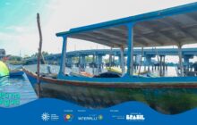 Fotografia horizontal em plano detalhe, capturando a proa de uma canoa grande e rústica atracada na água. A canoa tem o casco em tons de azul e verde desgastados e um teto baixo azul-claro com parte interna branca. Um mastro fino de madeira se eleva na proa. Em primeiro plano, a canoa está ligeiramente à direita do centro. No interior da canoa e em segundo plano, várias outras pequenas canoas de pesca coloridas (amarelo, vermelho, azul) estão atracadas, visíveis através da estrutura da primeira embarcação. O rio tem água calma e reflete levemente as cores do céu. Em terceiro plano, a cena é dominada por uma longa e alta ponte de concreto e vigas, que se estende horizontalmente sobre a água. Prédios distantes da cidade são visíveis sob a ponte. O céu é azul claro com nuvens esparsas. A cena contrasta a tradição caiçara das embarcações com a modernidade da infraestrutura urbana. O logo do projeto "Pá na Água" e a faixa azul dos apoiadores estão na base inferior.