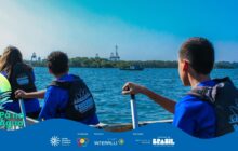 Fotografia horizontal em ângulo aberto, capturada da parte de trás de um pequena canoa de madeira. Três jovens, de costas, ocupam a embarcação. Eles usam camisas azuis e coletes salva-vidas pretos estampados com a palavra "Passaguá". Cada um segura um remo preto com cabo de metal. O jovem da extrema direita, em primeiro plano, é o mais próximo. Sua mão direita agarra o remo, que está fora d'água. À sua esquerda, um segundo jovem e, parcialmente visível à esquerda, uma terceira jovem. Todos estão olhando para o mar agitado. A água é de um azul profundo, com pequenas ondas. No segundo plano, sobre a água, há um pequeno barco verde, atracado e vazio. No horizonte, uma faixa de vegetação escura (mata ciliar) e, acima dela, gruas e guindastes portuários, indicando a proximidade de Paranaguá. O céu é azul-claro vibrante, quase sem nuvens, dominando o terço superior. Forte iluminação de dia ensolarado. Na parte inferior esquerda, em sobreposição, há o logo do projeto: duas pás de remo azuis com texto "Pá na Água". A imagem transmite ação e aprendizado prático no contexto caiçara.