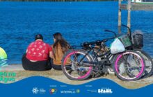 Fotografia horizontal em plano médio de duas pessoas sentadas de costas, relaxando à beira da água. O casal está sentado no chão em uma margem gramada, voltado para o corpo d'água azul escuro. À esquerda, um homem corpulento veste uma camiseta vermelha com estampa de palmeiras, boné preto e calça preta. À direita, uma mulher de cabelo longo e escuro usa uma camisa marrom-mostarda. Eles estão conversando, em atitude tranquila. Em primeiro plano, à direita, duas bicicletas pretas estão apoiadas na grama. A bicicleta mais próxima tem aro prateado e destaque de faixas e raios cor-de-rosa intenso nas rodas. Uma sacola plástica branca está pendurada no guidão da frente. A segunda bicicleta, parcialmente visível, está atrás. A água azul, profunda e agitada por pequenas marolas, domina o plano de fundo. Na extremidade direita superior, um pedaço de estrutura de madeira de um pequeno trapiche ou embarcação é visível. A iluminação é forte, de um dia ensolarado. Na base da imagem, a faixa azul com o logotipo do projeto "Pá na Água" aparece em sobreposição na lateral esquerda. A cena evoca um momento de descanso e contemplação.