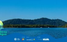 Fotografia horizontal em plano geral, dominada por uma paisagem natural composta por água, ilhas e céu. O corpo d’água, provavelmente o estuário de Paranaguá, ocupa a metade inferior, apresentando um azul escuro intenso com pequenas marolas. A luz solar forte reflete na superfície. No plano de fundo, erguem-se duas faixas de vegetação densa. A faixa mais próxima, no meio da composição, é de mata ciliar verde escura, com formações baixas de mangue e alguns pontos claros, onde se avistam 2 pequenos barcos brancos atracados ou ancorados. Atrás desta faixa, ao fundo, eleva-se uma grande montanha coberta por mata atlântica escura, que preenche toda a linha do horizonte. O céu é azul-claro, sem nuvens, ocupando o terço superior. A luz do dia é plena e intensa. Na lateral esquerda inferior, há o logo em sobreposição "Pá na Água", e na base, a faixa azul com os logotipos dos patrocinadores e realizadores do projeto. A cena é de vasta tranquilidade natural.