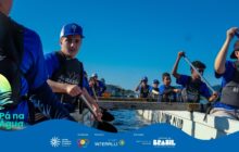 Fotografia horizontal em plano médio, capturada de dentro de uma canoa longa e branca. Aproximadamente 8 jovens, meninos e meninas, estão sentados em pares e engajados na remada. Todos usam coletes salva-vidas pretos sobre camisas azuis e bonés. O jovem em primeiro plano, no lado esquerdo, é o principal destaque. Ele usa boné branco e azul, olha diretamente para a câmera e segura o remo com as duas mãos, pronto para mergulhá-lo na água. À sua direita, apenas o braço e a camisa azul de outro participante são visíveis. [PAUSA BREVE] No centro, ao fundo, outros jovens remam com energia. O jovem na extrema direita, em plano mais próximo, também está ativo na remada, com o remo levantado acima da cabeça. A água azul escura é visível na lateral da canoa. No plano de fundo, sobre o horizonte, avista-se o perfil de uma pequena área urbana, com construções brancas e telhados claros, indicando a proximidade da cidade portuária. O céu é azul-claro intenso. A iluminação é forte, de dia ensolarado, realçando o branco do barco e o azul uniforme das camisas.