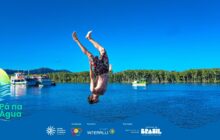Fotografia horizontal em plano aberto, capturando uma ação congelada. Um jovem, de tronco nu e shorts florais escuros, realiza um mergulho acrobático, de cabeça para baixo, no ar. Ele está no centro da imagem, com as pernas esticadas e os pés ligeiramente acima da cabeça, a cerca de 1 metro da água. O jovem é o foco nítido e ocupa o centro da cena contra o céu azul intenso, que preenche o terço superior. Em segundo plano, a água escura do rio reflete o céu. Na margem oposta, uma densa faixa de mata atlântica verde escura se estende horizontalmente, separando o céu e a água. Flutuando, à esquerda, há um barco de passageiros branco e amarelo e, à direita, um pequeno barco de pesca amarelo e verde. A luz solar é forte, criando um clima de liberdade e aventura. Na base, há uma faixa azul com o logo do projeto e os créditos de patrocínio.