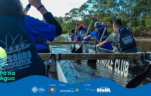 Fotografia horizontal em plano médio, vista de dentro de uma canoa de expedição lotada de jovens. A canoa, de madeira com laterais em preto e branco, tem a inscrição "PADDLE CLUB" visível em sua lateral. O primeiro plano é dominado pelas costas e ombros dos remadores, voltados para a frente. O jovem mais próximo, à esquerda, usa uma camisa azul com estampa branca e um colete salva-vidas preto, com o remo parcialmente visível. Os demais participantes estão alinhados em pares ao longo da embarcação, todos usando coletes salva-vidas e camisas azuis, remando ativamente. Os remos estão erguidos em ângulos variados, mergulhando na água escura do rio. A água está agitada pelo movimento da remada, com reflexos nas laterais internas do barco. No plano de fundo, a margem do rio é composta por uma faixa estreita de areia e terra e uma parede densa de mata e árvores com folhagem verde intensa. O céu não é visível devido ao enquadramento fechado na vegetação. A iluminação é de dia claro, filtrada pela copa das árvores. Na parte inferior esquerda, em sobreposição, o logo "Pá na Água" aparece.
