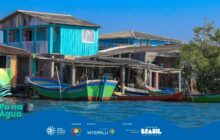 Fotografia horizontal em plano geral, exibindo casas simples e coloridas de uma comunidade ribeirinha, construídas em palafitas ou sobre a margem da água. A casa em destaque, à esquerda, é de um azul turquesa vibrante, com janelas brancas e um telhado escuro. Outras construções de cores azuis e brancas aparecem atrás. Em primeiro plano, uma fileira de canoas tradicionais de madeira está atracada na água, na frente das casas. As canoas são pintadas com cores fortes: a mais próxima é amarela e verde, a próxima é vermelha e verde-clima, e uma terceira, à direita, é verde e azul, com o nome "PARANAGUÁ" em destaque em algumas delas. A água, escura e calma, está no terço inferior, com pequenas marolas refletindo o céu. O céu, no terço superior, é de um azul intenso e claro, sem nuvens. A luz solar é plena, destacando a paleta de cores fortes do cenário cultural caiçara. Na parte inferior esquerda, em sobreposição, o logo "Pá na Água" aparece.