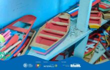 Fotografia em close-up de artesanato caiçara. O foco está em uma prateleira de madeira clara onde miniaturas de barcos estão dispostas. A cena é capturada em ângulo ligeiramente de cima para baixo. Em primeiro plano, no centro, repousa uma canoa de fundo bege com interior pintado de vermelho vivo, lateral azul-claro e detalhe rosa na parte inferior da proa. À esquerda, em outra canoa maior, vemos vários remos miniatura, vermelhos e um rosa com ponta verde e amarela, encostados na borda. Outras miniaturas estão dispostas em planos inferiores e superiores, parcialmente desfocadas. No canto superior direito, há mais dois barcos, um amarelo e um verde. O fundo é uma parede azul-claro. A iluminação é forte e artificial, realçando as cores vibrantes do artesanato. No canto inferior esquerdo da foto, o logotipo "Pá na Água" é sobreposto.