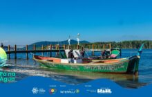 Fotografia horizontal de cena caiçara sob céu azul e claro. Em primeiro plano, no centro, está uma grande canoa de madeira, parcialmente atracada na areia clara. O barco é predominantemente verde, com detalhes em amarelo e a palavra "Paranaguá" pintada em verde-escuro na lateral. A proa e a popa têm detalhes em vermelho e verde-escuro. Três homens estão a bordo: um de camiseta branca na proa, um de camisa vermelha ao centro e outro de camisa marrom na popa. Todos usam bonés e parecem estar trabalhando no barco. Ao fundo, um píer de madeira estende-se sobre a água calma, com pilares escuros. No canto esquerdo do píer, uma pessoa está de pé. Na linha do horizonte, morros verde-escuros e uma densa vegetação nativa emolduram a água azul. A iluminação natural é intensa, criando reflexos na água e areia. A atmosfera é de trabalho e cotidiano ribeirinho.