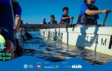 Fotografia horizontal e dinâmica em um barco de remo esportivo. A perspectiva é baixa, na altura da água escura, que ondula intensamente junto à lateral branca da embarcação. No barco, quatro jovens rapazes vestindo camisetas azuis e coletes salva-vidas pretos estão em ação de remada. Os rostos visíveis, da esquerda para a direita: um jovem no centro com expressão concentrada; outro jovem atrás dele, sorrindo; e um jovem em primeiro plano à direita com boné escuro, segurando o remo. O remo do jovem da direita entra na água. Mais à esquerda, apenas as mãos e braços de outro remador são visíveis, segurando o remo na vertical. A lateral do barco exibe os dizeres "TEAM MIRAGE CLUB" em letras pretas. O céu é azul-claro, sem nuvens. A iluminação é de sol forte, realçando o contraste entre o branco do barco e o azul escuro da água. Atmosfera de esporte e concentração.