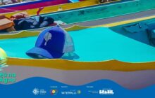 Fotografia close-up e horizontal, mostrando o detalhe do convés de um barco. Em primeiro plano, no centro, repousa um boné de trucker azul-marinho e tela branca. O boné tem o logo do projeto, uma pena e uma gota de água, em branco. O boné está sobre uma superfície azul-turquesa que é o convés do barco. Uma faixa amarela intensa contorna o convés, no terço inferior da imagem, servindo como linha guia. Em segundo plano, no canto superior direito, há uma peça de metal azul-turquesa, que é parte do casco do barco. Ao fundo e em desfoque, listras de outras embarcações atracadas trazem cores quentes como vermelho e amarelo. Uma almofada laranja dobrada e um tecido escuro aparecem no canto superior esquerdo. A iluminação é natural e direta, destacando o forte contraste entre o azul-turquesa, o amarelo e o azul do boné.