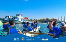 Fotografia horizontal em plano médio-aberto. A cena mostra um grupo de cinco jovens de costas, remando em uma canoa branca no rio ou baía. Todos usam coletes salva-vidas e camisetas azuis com o logo do projeto nas costas, e seguram remos com pás pretas. Eles estão voltados para o centro do quadro, com os remos erguidos, prontos para a remada. A água, azul escura e ondulada, preenche o primeiro plano. Ao fundo, à esquerda, dois barcos grandes de passageiros ou pesca estão ancorados: um azul e branco, e outro branco com detalhes em verde e vermelho. O horizonte é delimitado por uma faixa de palmeiras verdes. À direita, surgem estruturas brancas e pontiagudas de uma construção moderna, talvez um terminal marítimo. O céu, no terço superior, é de um azul intenso, sem nuvens. Na base, há uma faixa azul com o logo do projeto e os créditos de patrocínio.