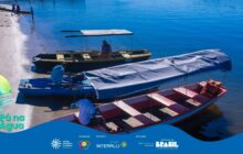 Fotografia horizontal em plano aberto. Três barcos de madeira de tamanhos variados atracados em uma margem de areia, em primeiro e segundo plano. A água do rio ou baía é azul escura, preenchendo a maior parte do lado direito. Da direita para a esquerda, os barcos são: um maior, vermelho e branco com toldo azul claro e coletes salva-vidas laranjas no interior; um médio, azul claro com toldo cinza; e um menor, amarelo e preto com toldo cinza. Três homens interagem com os barcos. Um, de camisa vermelha, está agachado no barco maior, perto da proa. Outro, de camisa azul, está sentado na lateral do barco médio. O terceiro, no barco amarelo, está sentado sob o toldo, remando. Ao fundo, no canto superior esquerdo, 3 pessoas em pé na areia e uma canoa atracada. A luz solar forte destaca o brilho da água e as cores vivas das embarcações.