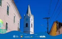Fotografia vertical do centro histórico, em plano de baixo para cima. A torre da Igreja de Nossa Senhora do Rosário, branca e alta, domina o centro, pontuando o céu azul intenso e sem nuvens. O campanário quadrado é branco, com quatro relógios pretos e ponteiro marcando aproximadamente 11:45h. No topo, um pináculo cinza claro e uma cruz preta. À esquerda da torre, um casarão colonial, de cor rosa pálido e com janelas verdes verticais. Sua fachada ocupa o terço esquerdo da imagem. À direita, parte de outro casarão com fachada amarela escura e um telhado cinza. Um poste de madeira, na extrema direita, sustenta linhas de energia escuras que cortam o terço superior. A forte iluminação solar destaca a brancura da torre, criando alto contraste com o céu azul.