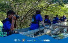 Fotografia horizontal registra jovens da oficina de fotografia navegando em um manguezal. A canoa estreita, cinza-clara, ocupa o centro da imagem. Na lateral da embarcação, lê-se "Mirage". Seis jovens, de costas ou em perfil, usam camisetas azuis vibrantes e coletes salva-vidas pretos. Em primeiro plano, à esquerda, um jovem de pele morena, cabelo escuro e colete preto, aponta uma câmera DSLR preta para frente, registrando a paisagem. No centro, o segundo jovem rema, com o remo parcialmente submerso na água escura. Mais quatro jovens e outros remos seguem à direita e ao fundo da canoa. A água do rio é calma e reflete a vegetação densa. O fundo e as laterais são dominados pelas raízes e galhos tortuosos do manguezal. A folhagem verde-escura cria um túnel natural sobre a água, conferindo uma atmosfera úmida e imersiva. Abaixo, logotipos institucionais.