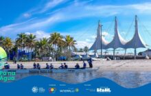 Fotografia horizontal em plano aberto. Uma canoa longa de cor branca, do "Passaúna Paddle Club", flutua próxima à margem, ocupando o centro da base. Dentro dela, seis jovens, com camisetas azuis e coletes salva-vidas escuros, estão sentados, segurando remos. Um sétimo jovem está em pé na popa, orientando. A canoa está voltada para a direita, parcialmente na água calma, que reflete o céu azul. A cena se passa em uma praia de areia clara e larga. No segundo plano, uma fileira de coqueiros altos e verdes se estende horizontalmente, separando a praia do céu. Logo atrás dos coqueiros, à direita, destaca-se uma estrutura arquitetônica moderna e branca, com três grandes "velas" de tecido tensionado, emoldurando o horizonte. Edificações brancas e escuras de um centro urbano aparecem no canto direito. O céu é de azul vibrante, com poucas nuvens brancas, sob luz intensa do sol. Logotipos institucionais aparecem no rodapé.