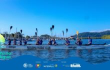 Plano médio horizontal registra uma grande canoa polinésia branca, inscrita com "Passaúna Paddle Club" no centro e "Team Mirage" nas laterais. Oito jovens, todos usando uniformes azuis e coletes salva-vidas escuros, estão sentados na canoa. Eles erguem seus remos pretos em sincronia acima da cabeça, em um gesto de celebração. Um dos remos é amarelo e se destaca à direita. A canoa navega em água azul escura e levemente agitada, que preenche o primeiro plano. No segundo plano, atrás dos remadores, vê-se um horizonte costeiro. À esquerda, mastros de veleiros atracados e estruturas de uma marina; ao centro-direita, uma embarcação maior branca; e à direita, a silhueta de morros cobertos por mata nativa verde-escura. O céu é azul claro, sob forte luz solar. A cena transmite energia e união da equipe. Logotipos institucionais aparecem no rodapé.