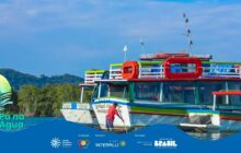 Plano médio de uma embarcação de passageiros atracada na água escura, exibindo cores vibrantes. O barco tem casco branco e dois andares superiores com parapeitos em verde limão, azul e vermelho. Janelas grandes, de formato quadrado e retangular, pontuam a lateral branca. Na proa, o nome da embarcação, parcialmente visível, inicia com "Senhor Manuel An[...]", pintado em azul. Em primeiro plano, um homem de pele morena, camiseta vermelha escura e calça preta, está de costas, curvado, limpando a lateral do barco com uma vassoura de cabo longo. Ele está com os pés próximos à borda da água. O segundo plano, à esquerda, é dominado por uma densa vegetação de manguezal de um verde-amarelado. Morros cobertos de mata verde-escura formam o horizonte ao fundo, sob um céu azul claro e sem nuvens. Logotipos institucionais aparecem no rodapé.