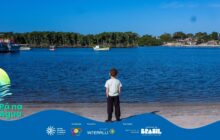 Fotografia em plano aberto e perspectiva frontal. Um vasto espelho de água azul escura e ondulada ocupa o centro do quadro. Centralizado na base, um menino de pele clara, cabelo castanho-claro e camiseta azul-clara (verde-água) está de costas, com as mãos cruzadas atrás do corpo. Ele usa calças ou bermudas escuras e tênis pretos e parece observar a paisagem aquática à sua frente. A criança está sobre uma estreita faixa de areia úmida e escura, em primeiro plano. Na linha do horizonte, o fundo é preenchido por uma faixa de vegetação densa e verde escura que se estende sobre a água, com árvores de mangue e restinga. À esquerda, barcos coloridos estão ancorados. No canto superior direito, um pequeno vilarejo de casas simples de cor clara e telhados escuros aparece na margem. O céu é de um azul intenso e sem nuvens. Logotipos institucionais aparecem no rodapé.