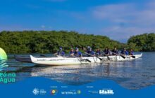 Fotografia horizontal em plano médio. Uma canoa polinésia (modelo OC6), branca e alongada, flutua sobre água escura, calma e reflexiva. Dez remadores, 8 homens e 2 mulheres, estão sentados em pares, perfilados. Todos vestem camisas azuis-escuras, bonés e remam com pás brancas, voltados para a direita. Na lateral da canoa, lê-se "TEAM MIPE" e "PARANAGUÁ PADDLE CLUB". A proa e a popa são escuras, e o barco tem um ama (flutuador lateral) branco. Em primeiro plano, no canto inferior esquerdo, uma pena estilizada verde e amarela sobre a água indica o nome do projeto: “Pá na Água”. Em segundo plano, uma densa e exuberante floresta de mangue verde-escura forma um paredão vegetal, ocupando mais da metade superior da imagem. Ao fundo, o céu é azul claro, com poucas nuvens, indicando tempo ensolarado e luz intensa. A composição foca na atividade de remo e no trabalho de equipe, integrada ao ecossistema local. Logotipos institucionais aparecem no rodapé.