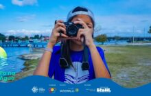 Fotografia horizontal em plano próximo, centralizada no busto de uma jovem fotógrafa. A jovem, de cabelo castanho-escuro e pele clara, usa uma camiseta azul-escura com um logotipo branco no peito e um boné cinza virado para trás. Ela segura uma câmera preta tipo mirrorless ou compacta avançada, cobrindo a maior parte do rosto. A alça preta da câmera está pendurada no pescoço. Ela tem pulseiras brancas e um colar com pingente prateado. A jovem está em uma área de terra e grama, na margem da água. O fundo está desfocado. À esquerda, vê-se a água azul escura do rio e, à direita, uma embarcação de pesca branca e azul está ancorada próximo a um pequeno cais. No plano de fundo, há um píer de concreto e metal, com árvores e algumas construções urbanas. O céu é azul claro, com nuvens brancas e suaves, indicando luz diurna e suave. A imagem foca na ação de registrar o ambiente. Logotipos institucionais aparecem no rodapé.